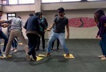 A group of teenagers doing an icebreaker exercise in which one person is blindfolded and the others are helping him jump across yellow tiles that are scattered across the floor